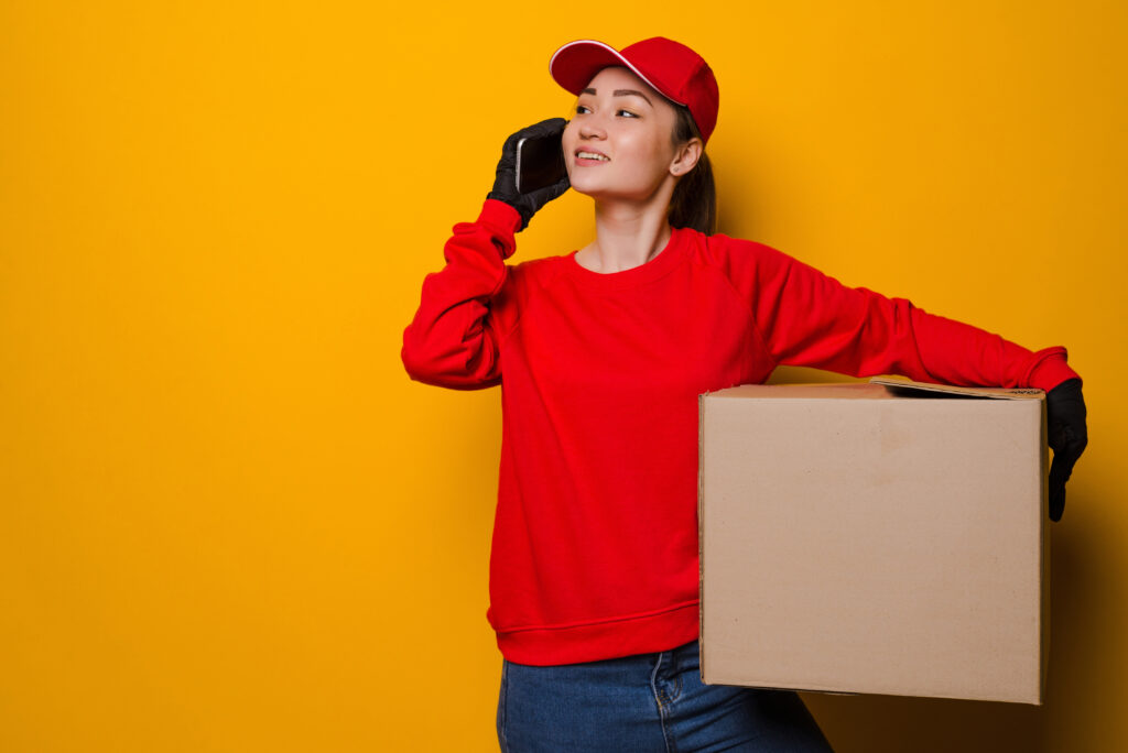 young asian delivery woman holding box talking on the phone isolated on yellow background